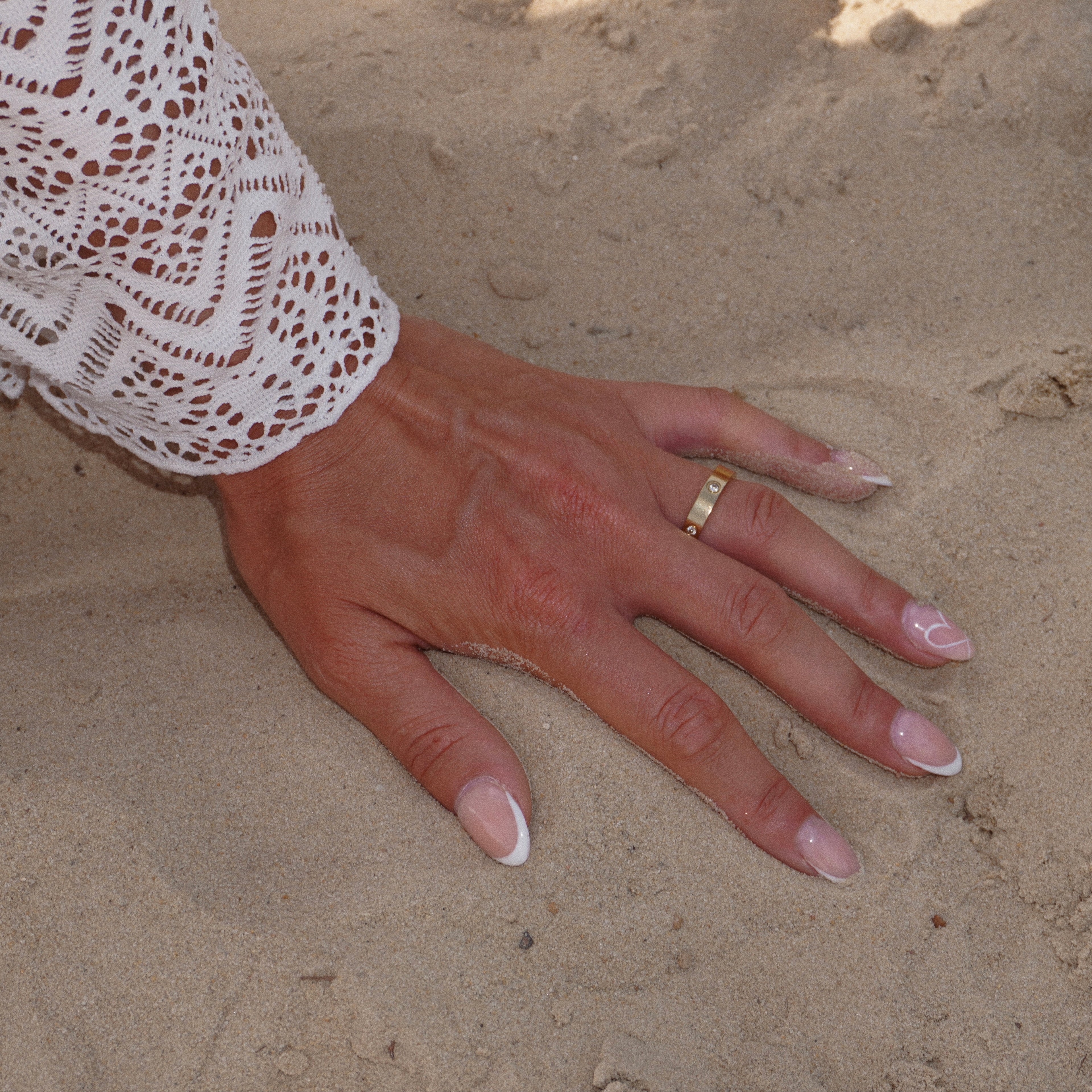 Hand im feinen Sand mit schlichtem, 18k vergoldetem vergoldetem Edelstahlring, besetzt mit runden Zirkonia-Steinen, minimalistischem Design. Eleganter Sommer-Look mit natürlichem Strand-Feeling, ideal kombinierbar mit anderen Goldschmuckstücken.