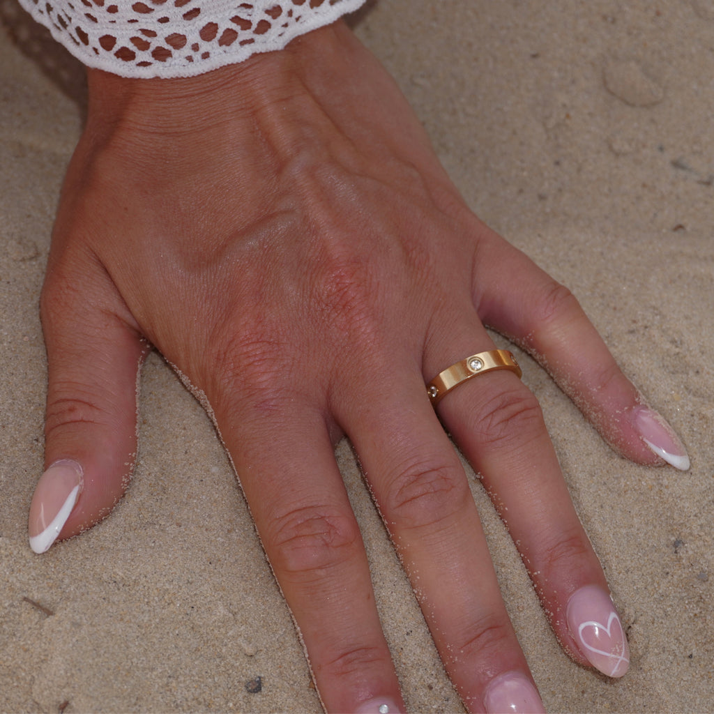 Hand im Sand mit breitem eleganten 18k vergoldetem Edelstahlring, besetzt mit Zirkonia-Steinen. Robustes, glänzendes Design. Sommerlicher Strandlook mit luxuriösem Goldschmuck.