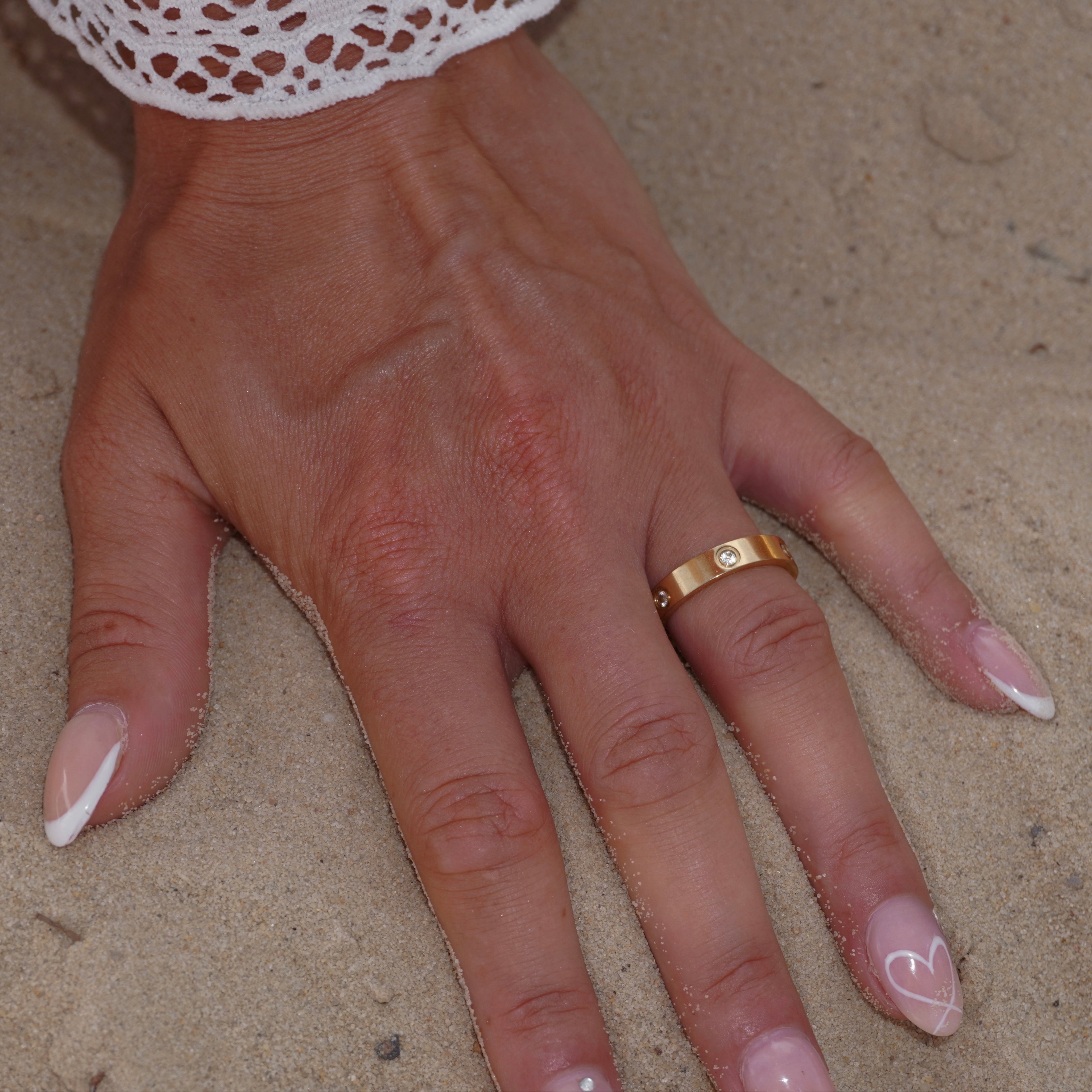 Hand im Sand mit breitem eleganten 18k vergoldetem Edelstahlring, besetzt mit Zirkonia-Steinen. Robustes, glänzendes Design. Sommerlicher Strandlook mit luxuriösem Goldschmuck.