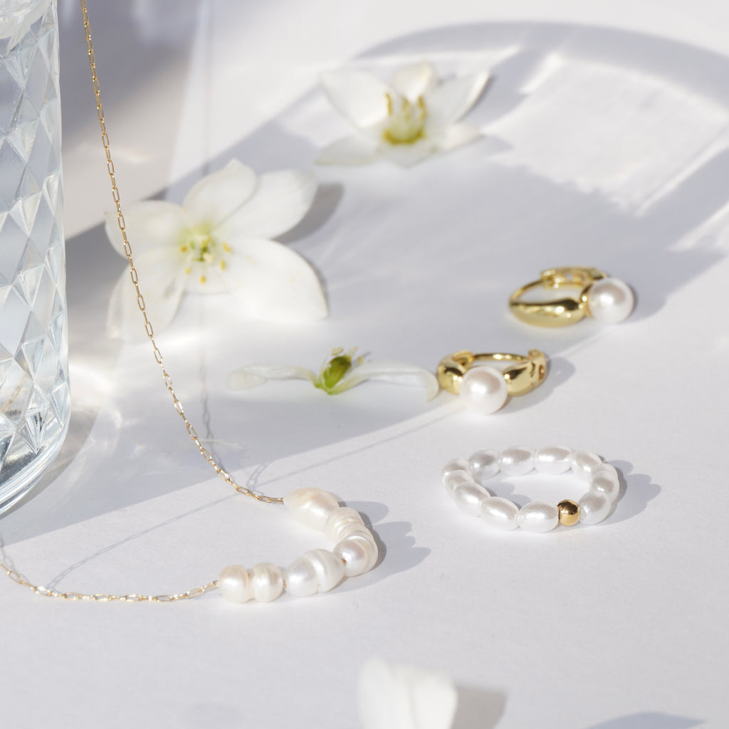 Perlenring aus simulierten weißen Perlen mit mittiger 14-karätig vergoldeter Edelstahl-Perle kombiniert mit goldenen Ringen und einer goldenen Süßwasserperlen Halskette, dekorativ auf weißer Fläche mit Blüten arrangiert. 
