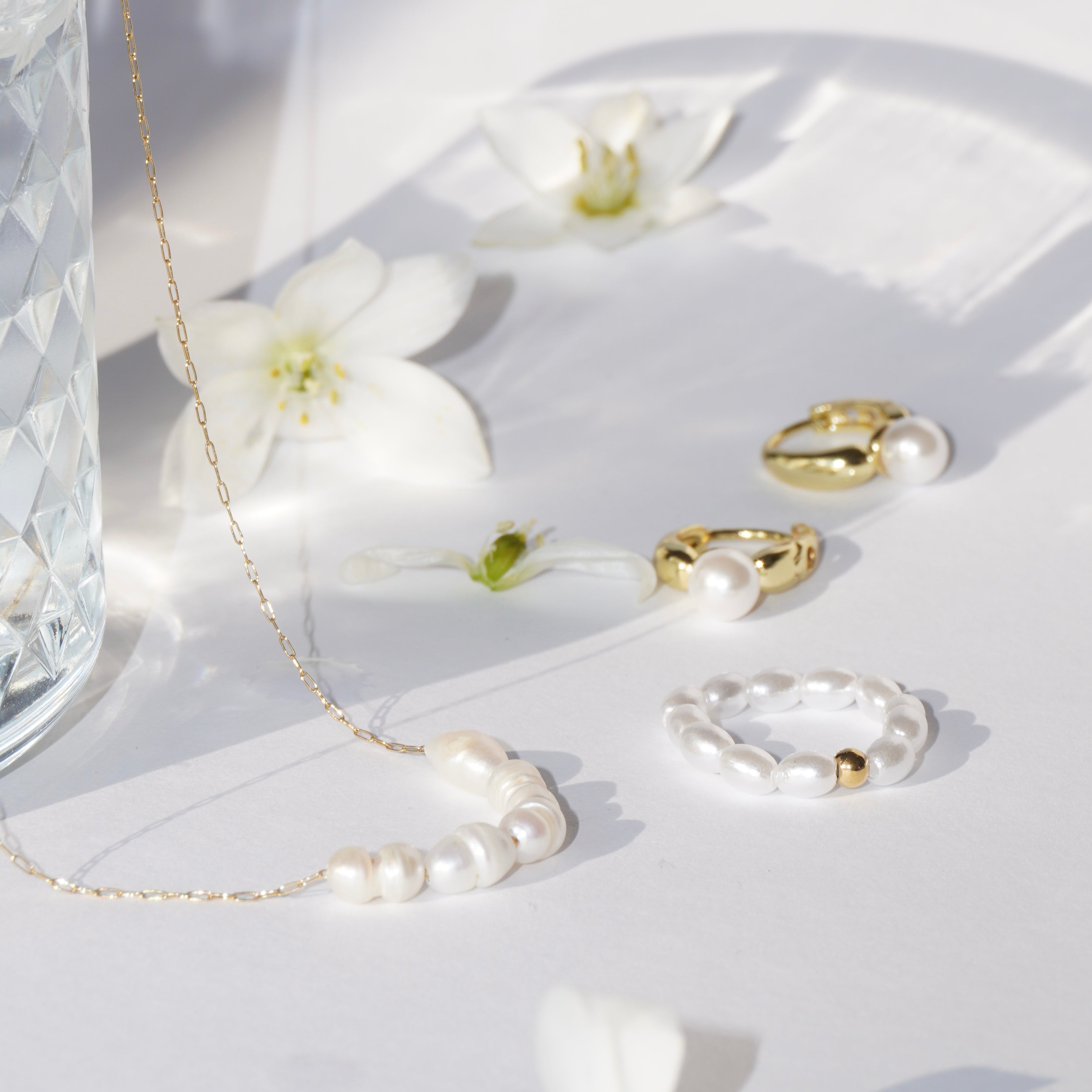 Perlenring aus simulierten weißen Perlen mit mittiger 14-karätig vergoldeter Edelstahl-Perle kombiniert mit goldenen Ringen und einer goldenen Süßwasserperlen Halskette, dekorativ auf weißer Fläche mit Blüten arrangiert. 