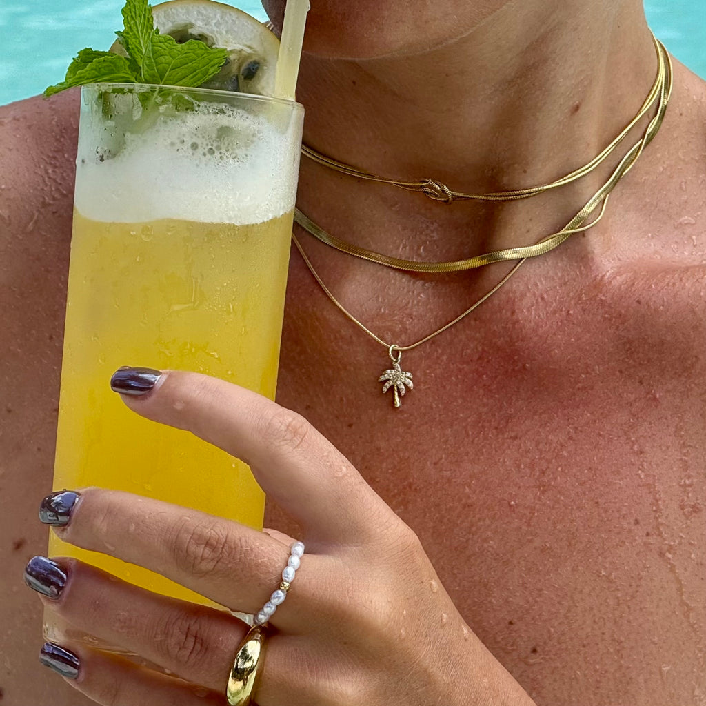 Frau im Pool mit zwei vergoldeten Edelstahl im Layering Look. Eine Schlangenkette und und schlichte Kette mit kleinem Palmen-Anhänger. Dazu 14k vergoldeter Edelstahl Ring und am Mittelfinger einen Perlenring aus simulierten weissen Perlen und einer 14-karätig vergoldeten Perle in der Mitte. Sie hält ein Glas mit gelbem Cocktail, Minze und Limette.