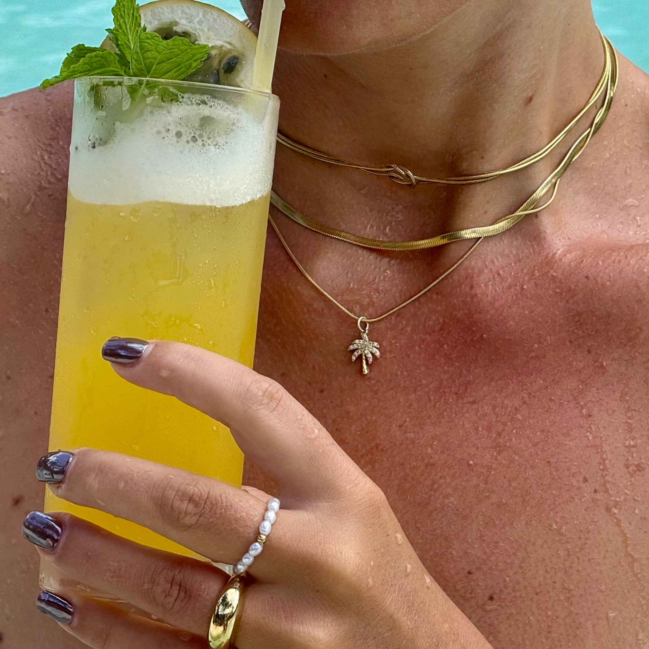 Frau im Pool mit zwei vergoldeten Edelstahl im Layering Look. Eine Schlangenkette und und schlichte Kette mit kleinem Palmen-Anhänger. Dazu 14k vergoldeter Edelstahl Ring und am Mittelfinger einen Perlenring aus simulierten weissen Perlen und einer 14-karätig vergoldeten Perle in der Mitte. Sie hält ein Glas mit gelbem Cocktail, Minze und Limette.