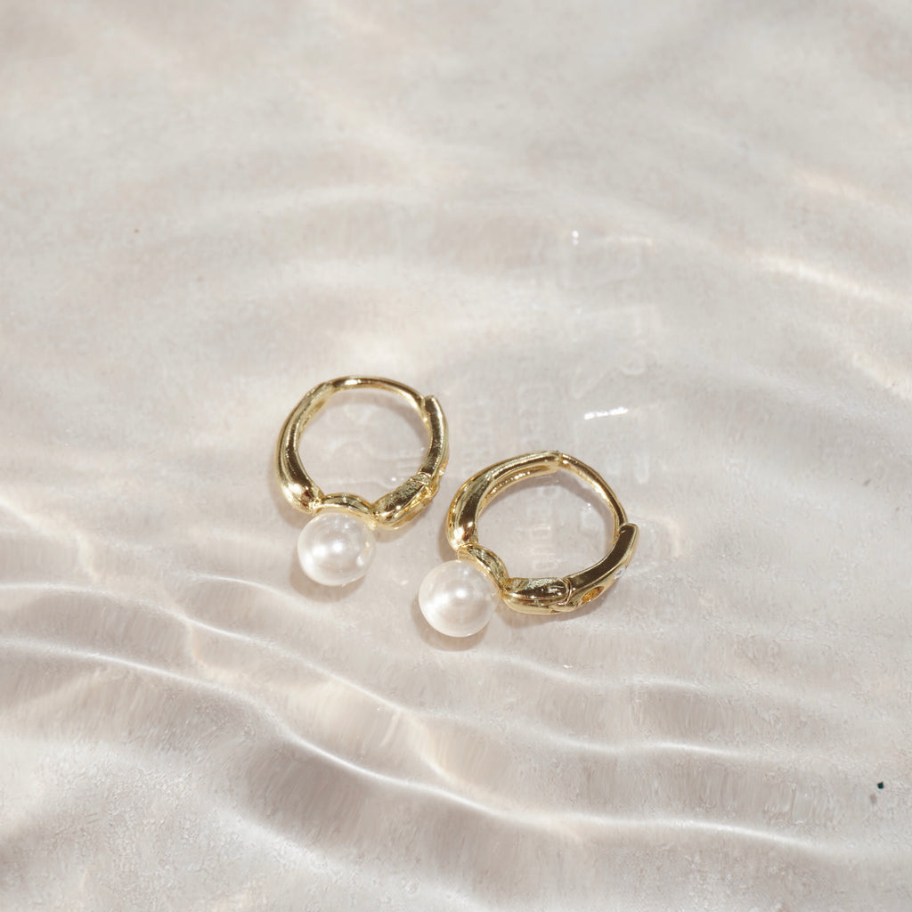 Zwei goldene Creolen mit Perlenanhänger, fotografiert im Wasser mit Sonnenreflexen.