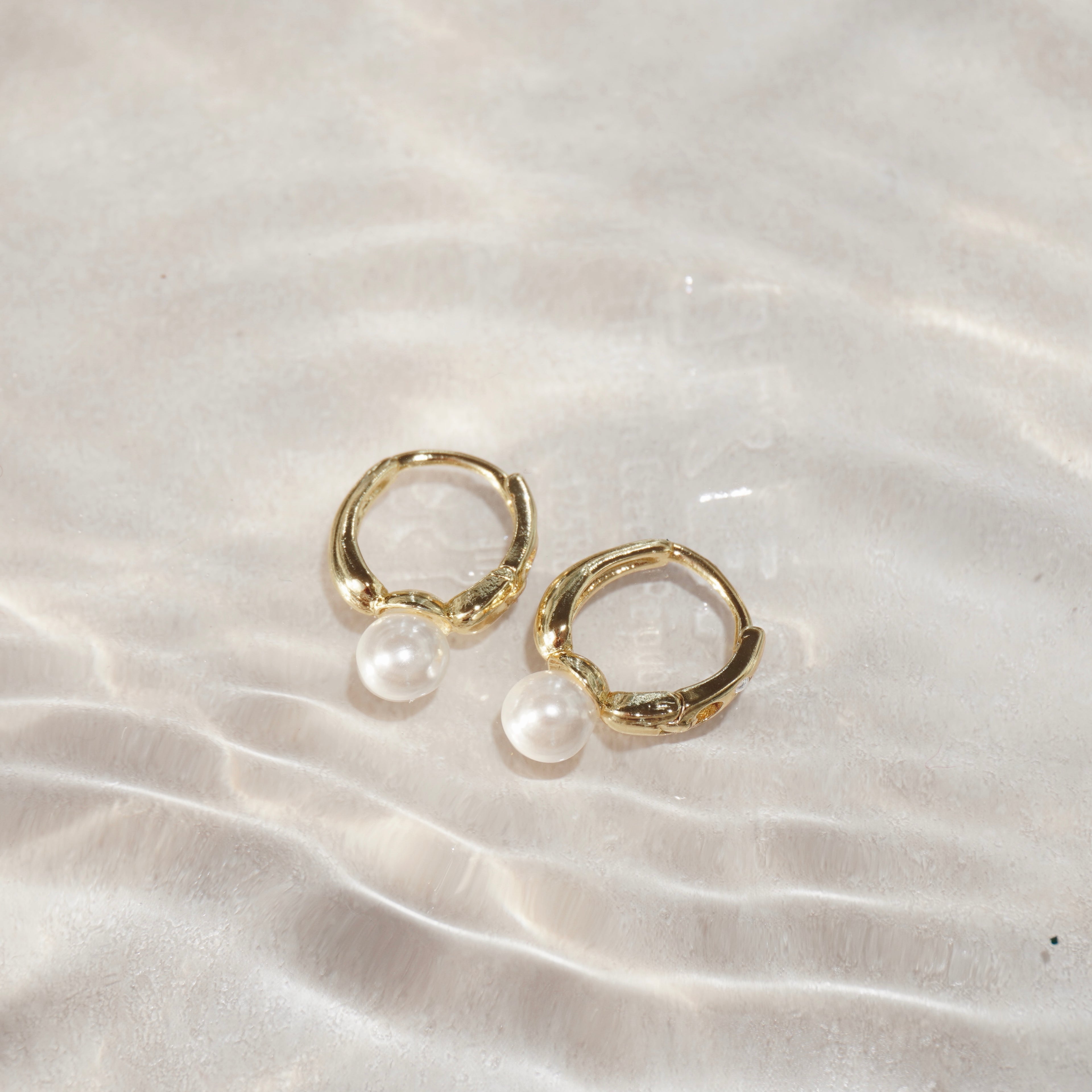 Zwei goldene Creolen mit Perlenanhänger, fotografiert im Wasser mit Sonnenreflexen.