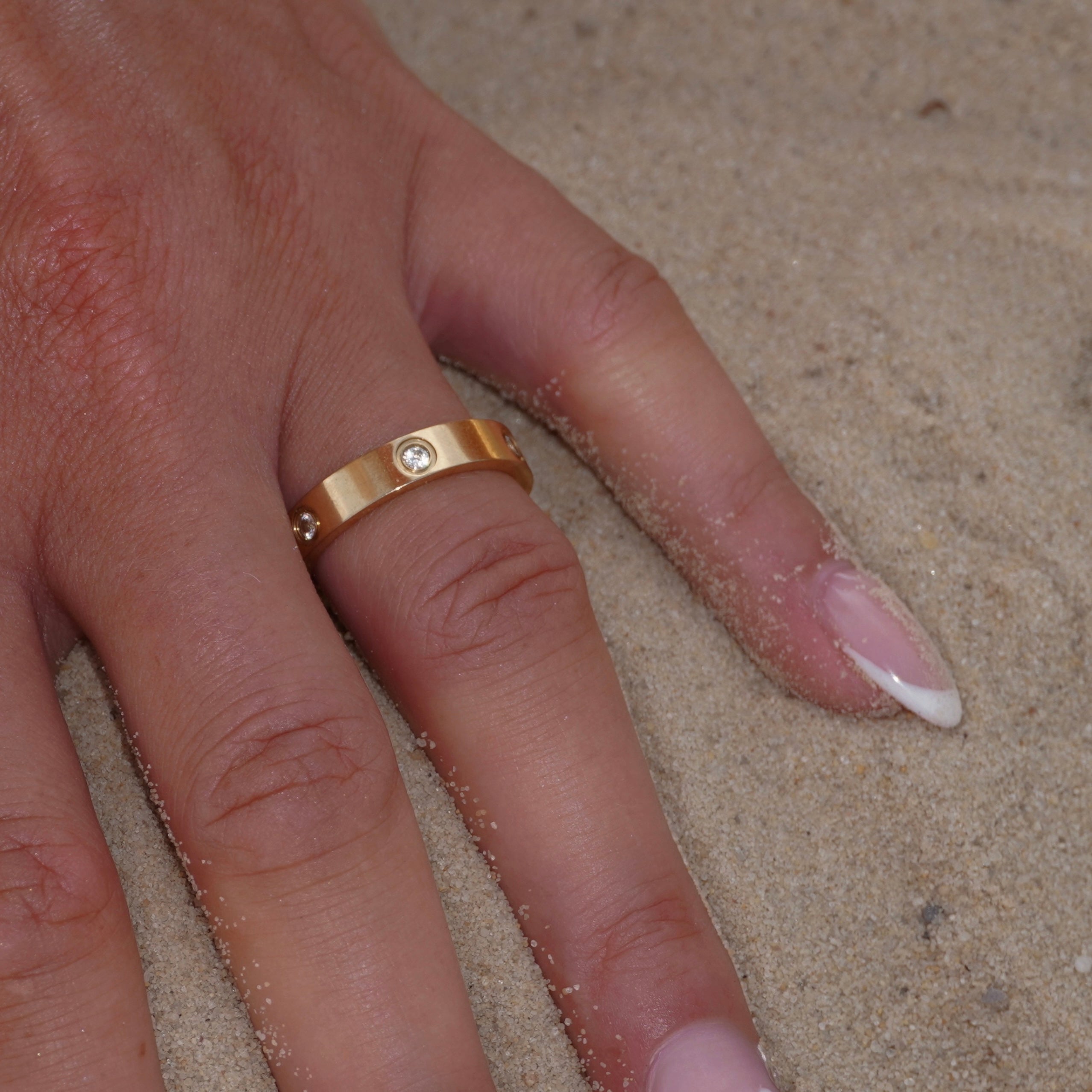Nahaufnahme einer Hand im Sand mit 18k vergoldetem Edelstahlring, verziert mit funkelndem Zirkonia-Steinchen. Hochwertiger und zeitloser Schmuck für elegante Outfits. 