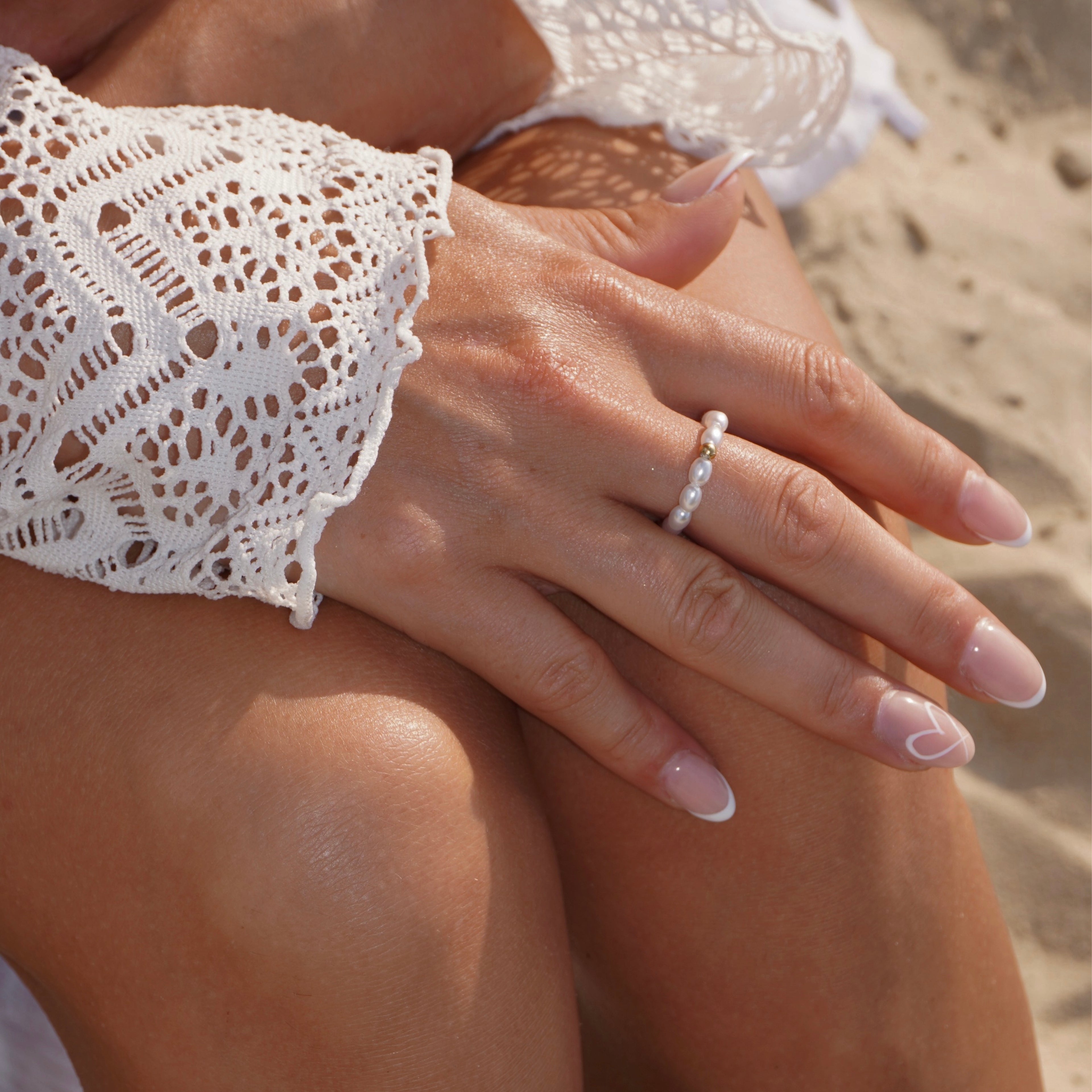Detailaufnahme einer Hand mit Perlenring aus simulierten weißen Perlen und einer mittigen 14-karätig vergoldeten Edelstahl-Perle, kombiniert mit natürlicher French-Maniküre, am Strand getragen.