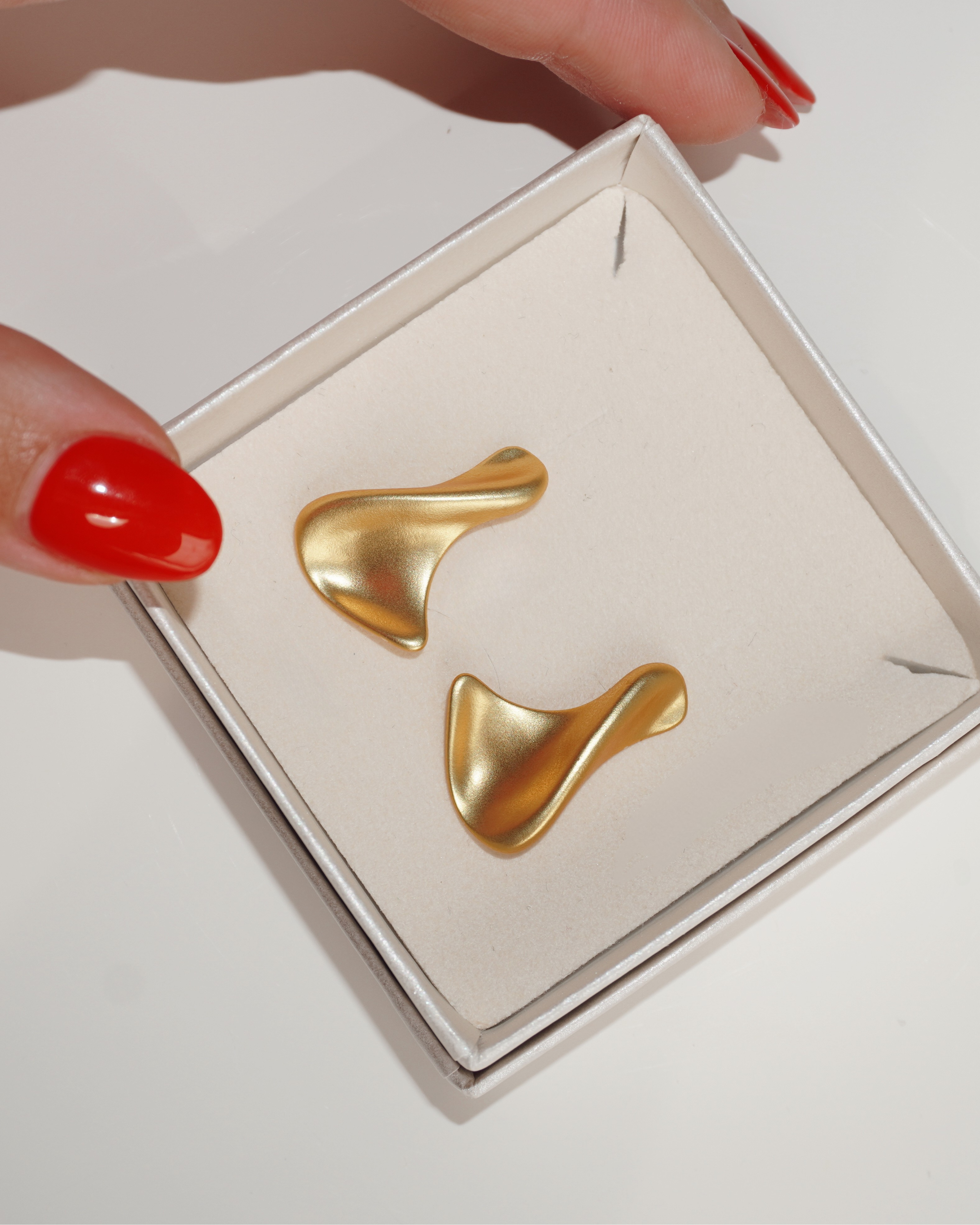 18k vergoldete O-Ringe aus 925er-Sterling Silber, leicht wellenförmig, liegend in einer Schmuckbox, mit rotenFingernägeln als Hintergrund