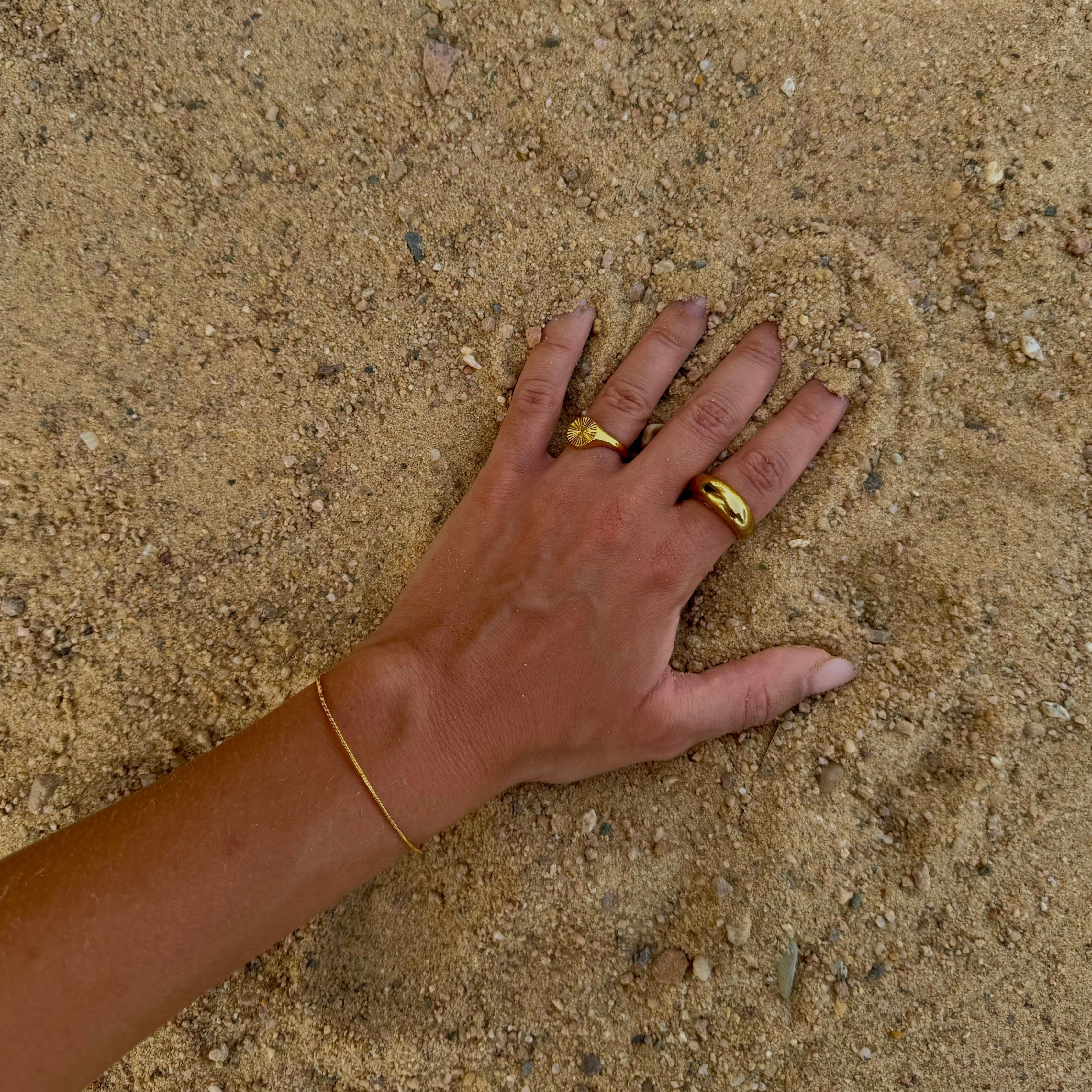Hand im Sand mit zwei goldenen 14k vergoldeten Edelstahlringen. Ring aus Sonnenoptik am Ringfinger und robuster Ring am Zeigefinger. Dazu ein zartes 14k vergoldetes Edelstahlarmband kombiniert. Minimalistischer und edler Strandlook mit Goldschmuck. 