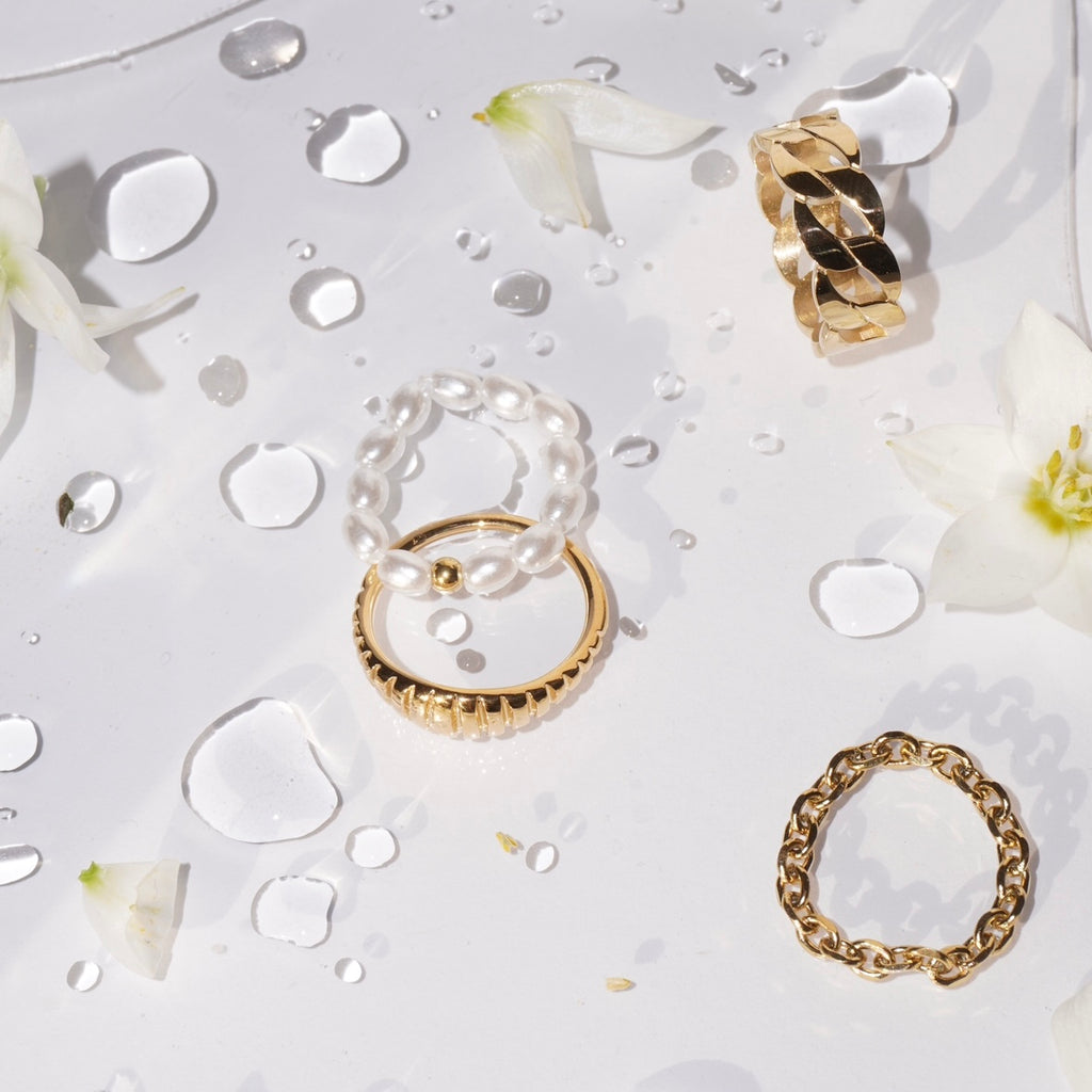 Flachliegende Aufnahme eines Perlenrings aus simulierten weißen Perlen und einer mittigen 14-karätig vergoldeten Edelstahl-Perle, daneben goldene Ringe, auf weißer Oberfläche mit Wassertropfen und weißen Blütenblättern. 