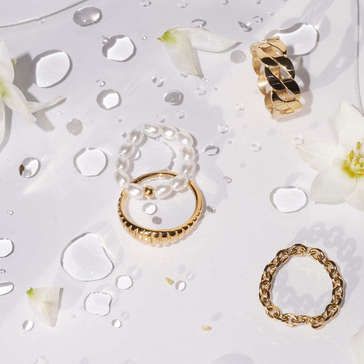 Flachliegende Aufnahme eines Perlenrings aus simulierten weißen Perlen und einer mittigen 14-karätig vergoldeten Edelstahl-Perle, daneben goldene Ringe, auf weißer Oberfläche mit Wassertropfen und weißen Blütenblättern. 